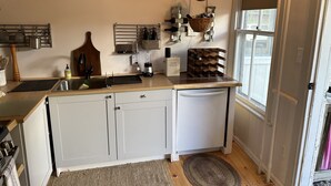 Une cuisine avec des meubles blancs, un lave-vaisselle, un évier et une fenêtre. On y trouve également des paniers suspendus et un porte-bouteilles fixé au mur.