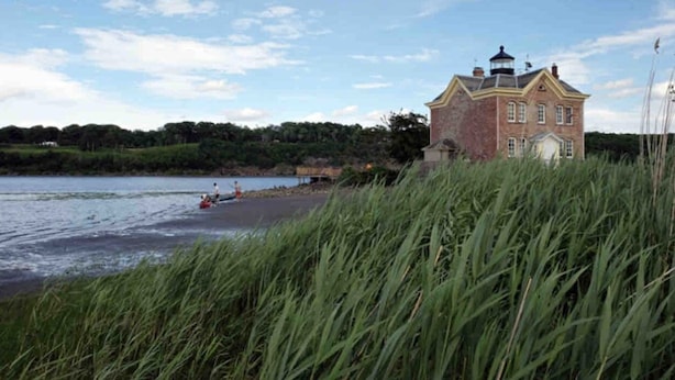 Un grand bâtiment en briques surmonté d’un phare, situé au bord d’une étendue d’eau, avec de hautes herbes au premier plan.