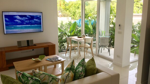 A modern living room with a flat-screen TV, wooden furniture, and a view of a lush garden through large sliding glass doors. - Private Garden Condo - 5 min from the Beach (Koh Samui)