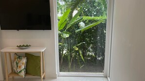 A bedroom with a bed, a TV mounted on the wall, a small table with a bowl and a pillow, and a sliding glass door leading to a garden. - Private Garden Condo - 5 min from the Beach (Koh Samui)