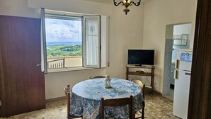 A room with a round table, chairs, a television, a refrigerator, and a window with a view of greenery. - S320 - Sirolo, one bedroom apartment with breathtaking view (La Madonnina)