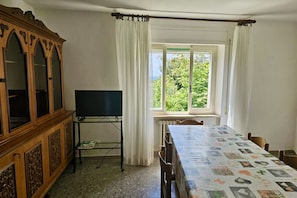 A room with a wooden cabinet, a television on a stand, a table with a quilt, and a window with curtains.