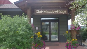 The entrance to Club Meadow Ridge with a wooden facade, glass doors, and a welcoming porch area with potted plants and flowers. - Dog-friendly condo close to Winter Park shuttle with pool, hot tub, tennis, gym (Fraser)