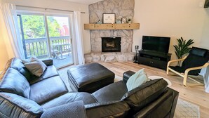 A living room with a stone fireplace, a black leather sofa, a TV, and a balcony with a view of greenery. - Dog-friendly condo close to Winter Park shuttle with pool, hot tub, tennis, gym (Fraser)