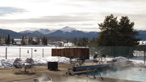 A pool area with chairs and a hot tub, surrounded by a wooden deck and a metal fence, with mountains in the background. - Dog-friendly condo close to Winter Park shuttle with pool, hot tub, tennis, gym (Fraser)