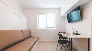 Une petite pièce moderne, dotée d’un canapé, d’une table à manger avec des chaises, d’un téléviseur fixé au mur et d’une fenêtre laissant entrer la lumière naturelle.