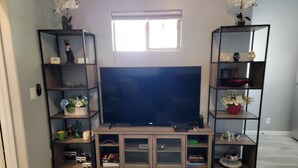 A living room with a large flat-screen TV on a wooden stand, flanked by two black shelving units with various decorative items and plants.