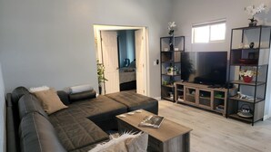 A modern living room with a sectional sofa, a coffee table, and a TV stand with a flat-screen TV. There is a bookshelf with decorative items and a door leading to another room.