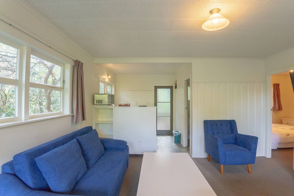 Un espace de vie compact comprenant un canapé bleu, un fauteuil assorti, un petit coin cuisine et une porte donnant sur une autre pièce.