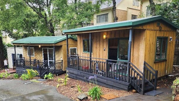 A row of wooden cabins with green roofs and black railings, each with a small porch and a window. - Touchwood Motor Inn (Auckland)