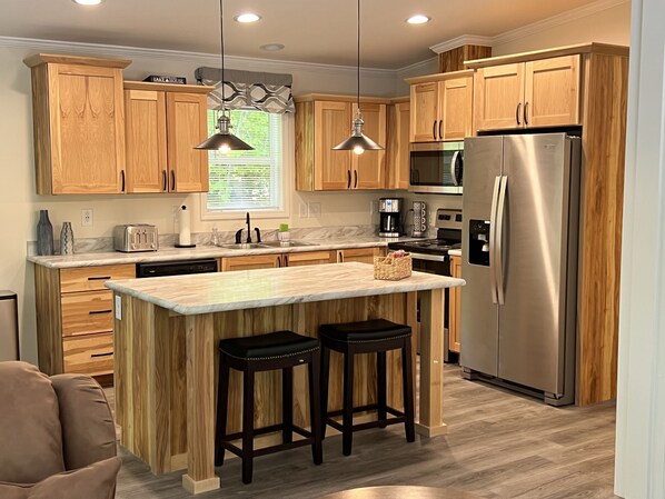 A modern kitchen with wooden cabinets, stainless steel appliances, a marble countertop, and two bar stools.