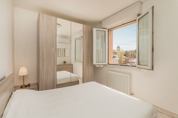 A hotel room with a bed, a nightstand with a lamp, a mirror, a window with curtains, and a radiator.