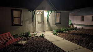 Une maison avec une porte d’entrée, deux fenêtres, un banc et un chemin menant à la porte.
