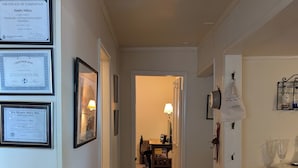 Un couloir avec des certificats encadrés accrochés au mur, un réfrigérateur, un panier rempli de fruits et une table avec une lampe.