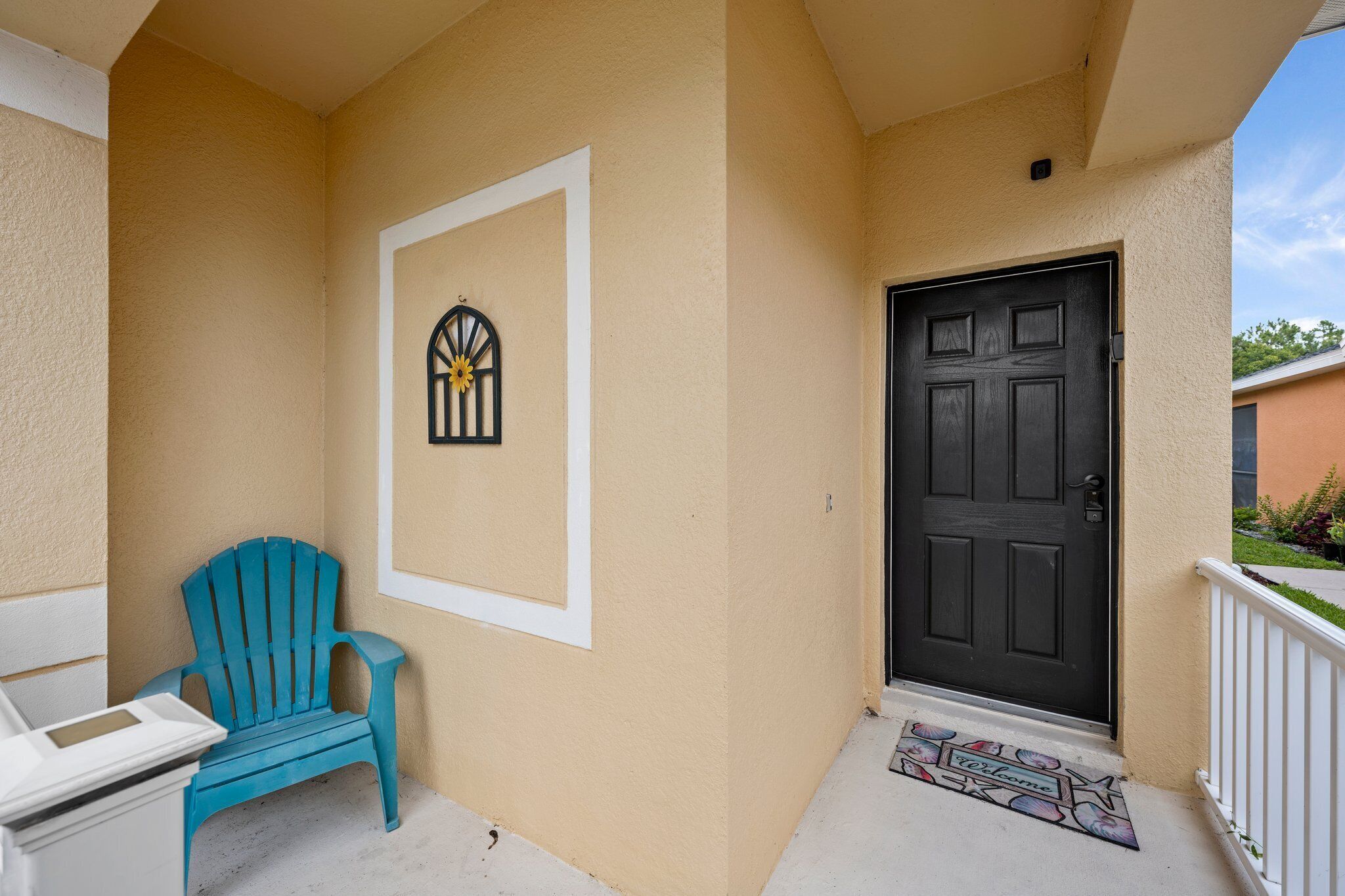 A beige exterior wall with a black door, a blue Adirondack chair, and a welcome mat.