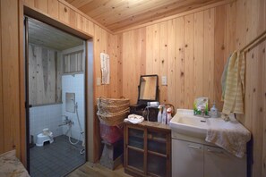 Un baño revestido de paneles de madera con inodoro, lavamanos y espejo. Hay una cesta y un pequeño armario con artículos de aseo.
