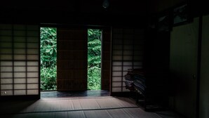 Ein traditioneller japanischer Raum mit Tatami-Böden, einer Schiebetür, die zu einem Garten führt, und einem Stapel gefalteter Matten rechts.