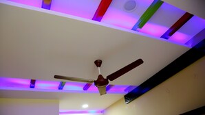 A ceiling fan with a light fixture and a recessed ceiling with colorful LED lighting. - DIVYA LODGE & HOTEL ROOMS (Hyderabad)