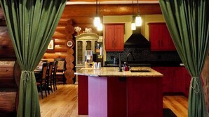A log cabin interior with a wooden floor, a central island with a sink, and a kitchen area with red cabinets and a black range hood. - The Lodge at Lakeshore (Emeryville)
