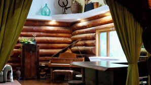 A log cabin interior with a wooden floor, a dining area with stools, a kitchen with a hanging pot rack, and a window with curtains. - The Lodge at Lakeshore (Emeryville)