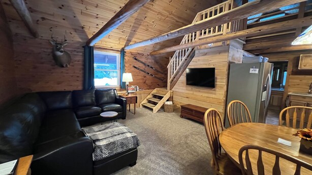 Een gezellig interieur van een chalet, met een zwarte leren bank, een houten eettafel met stoelen, een trap en een bevestigde televisie.