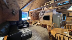 A cozy cabin interior with a black leather sofa, a wooden dining table with chairs, a staircase, and a mounted television. - Waterfall Cabin - Pictured Rocks Lodging (Munising)