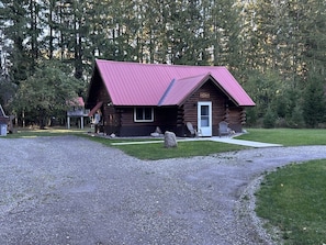 Uma cabana de tronco com telhado vermelho, uma porta da frente e duas cadeiras Adirondack na área externa.