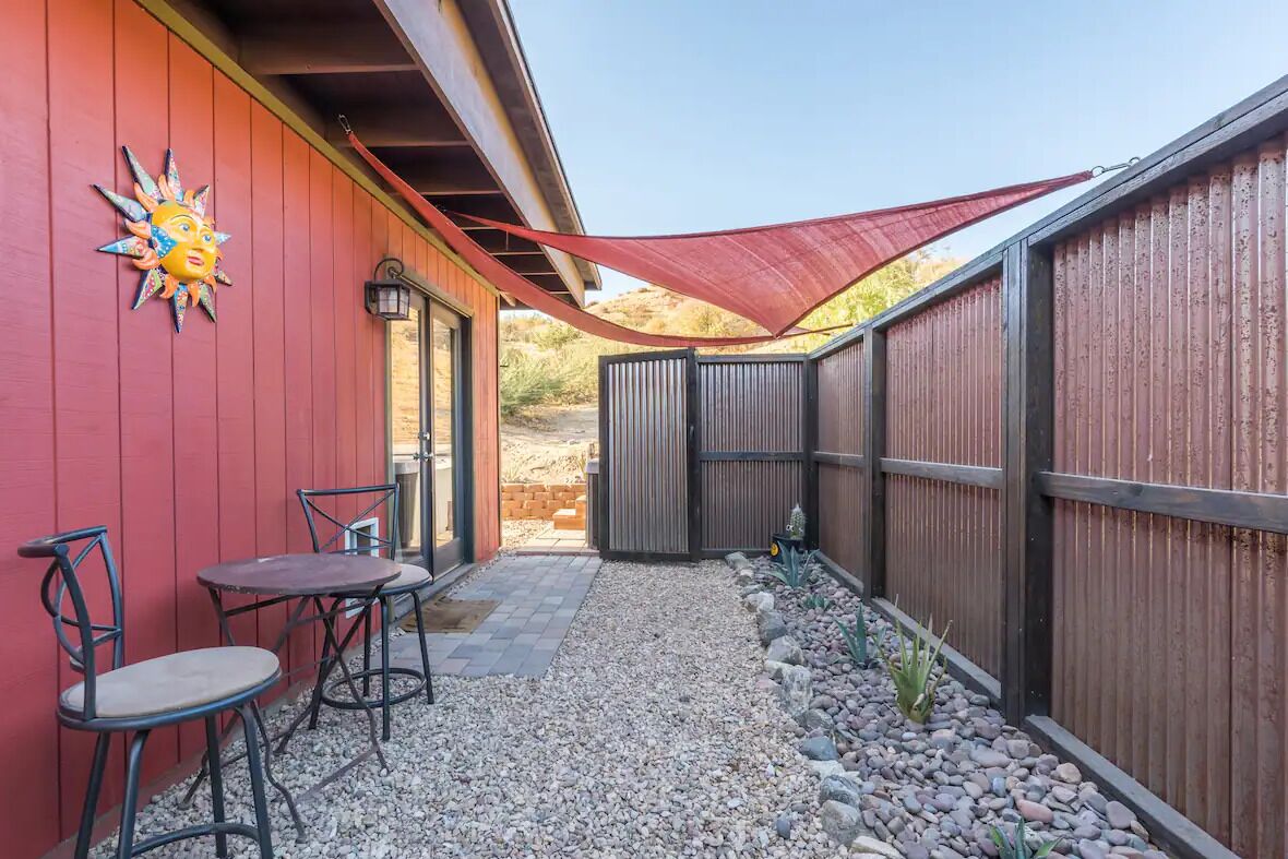 A patio with two metal chairs and a table, a red wall with a sun decoration, and a wooden fence.