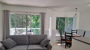 Un salon moderne avec un canapé gris, une table à manger et des chaises, ainsi que de grandes fenêtres donnant sur un paysage verdoyant.