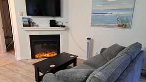 Un salon avec une cheminée, un téléviseur à écran plat fixé au-dessus, un canapé gris et un tableau représentant une scène de plage.