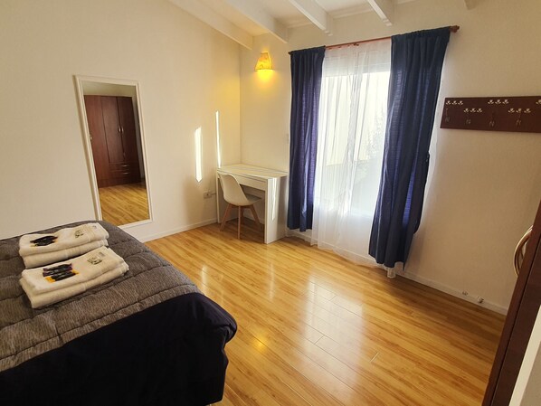 A bedroom with a bed, a desk, a chair, a mirror, and a window with curtains.