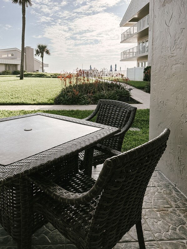 Een terras buiten met een tafel en rieten stoelen, een gazon en een gebouw op de achtergrond.