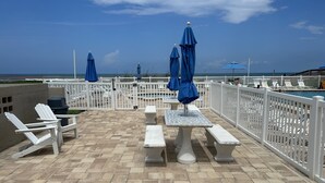 Un espace patio avec des chaises Adirondack blanches, une table surmontée d’un parasol bleu et une piscine en arrière-plan.