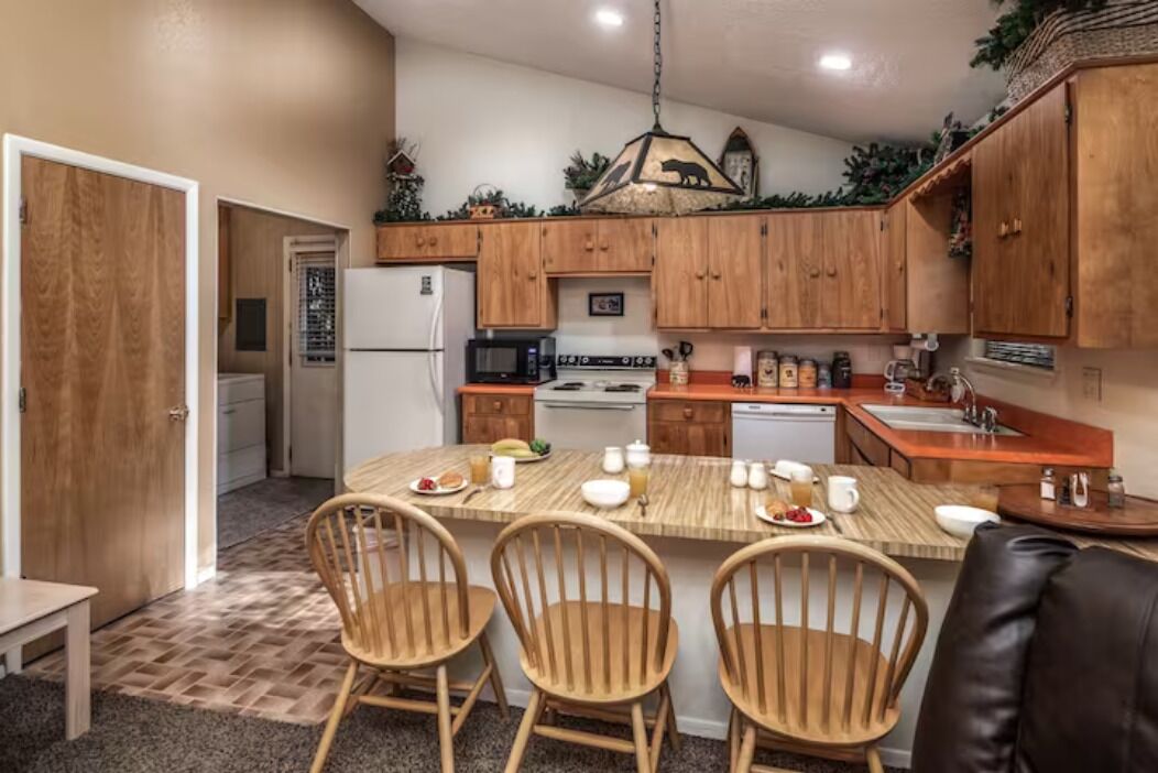 A compact kitchen with wooden cabinets, a central island with chairs, a refrigerator, microwave, stove, and sink.