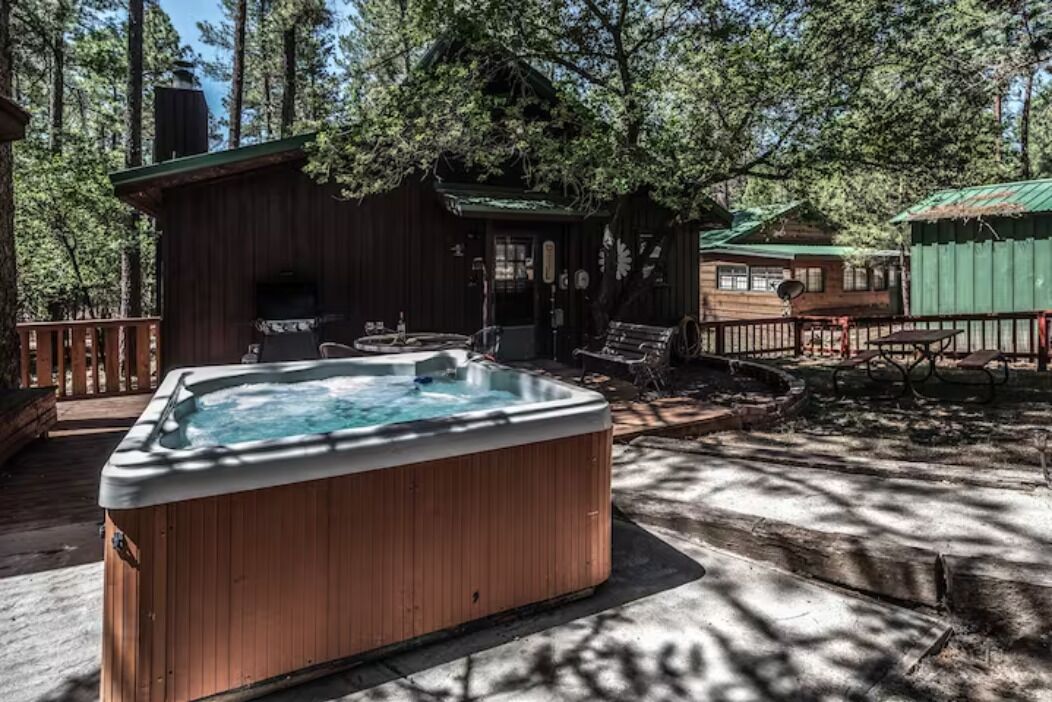 A wooden hot tub in a forested area with a cabin and outdoor furniture.