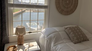 Ett sovrum med en säng, ett fönster med utsikt över en sjö eller ett vattendrag och en brygga, en vävd väggdekoration och ett sängbord med en lampa och en korg.