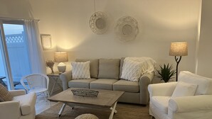 Ett vardagsrum med en beige soffa, vita fåtöljer, ett soffbord och två vägglampor.