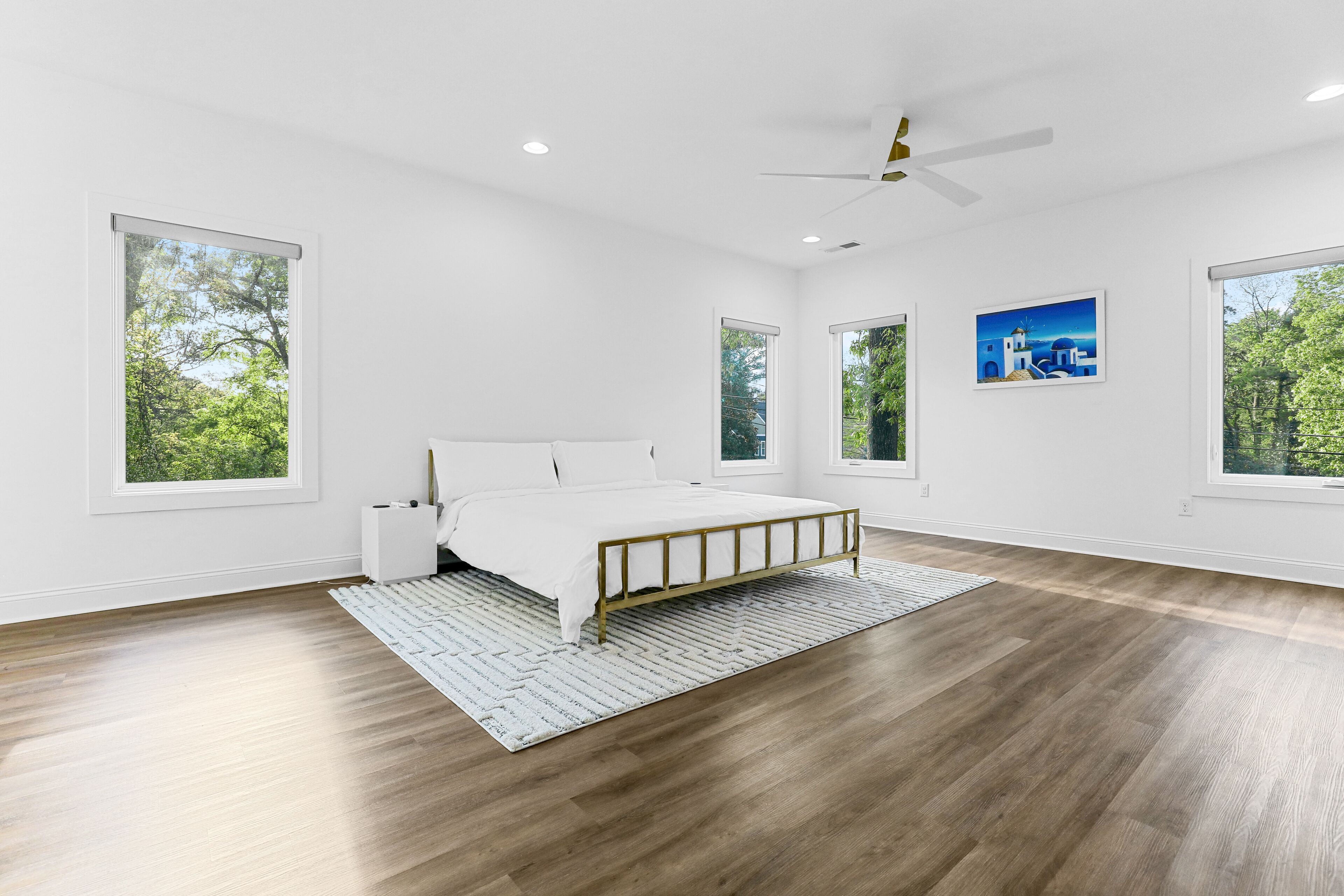 A spacious bedroom with a large bed, a ceiling fan, and multiple windows offering views of greenery.