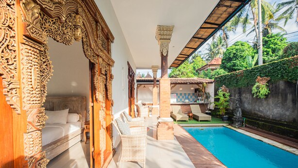 An indoor swimming pool area with a wooden bench and a sofa, surrounded by ornate wooden carvings and a view of the outside. - Supala Private Living B (Ubud)