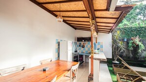 A spacious dining area with a wooden table and chairs, a kitchen in the background, and a small outdoor seating area with wicker chairs. - Supala Private Living B (Ubud)