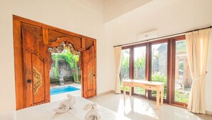A spacious room with a large bed, a wooden door with intricate carvings, a small wooden table, and a view of a swimming pool and greenery outside. - Supala Private Living B (Ubud)
