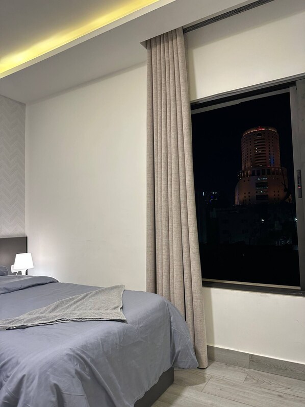 Une chambre d’hôtel moderne avec un grand lit, une table de chevet, une lampe et une fenêtre donnant sur une vue panoramique de la ville la nuit.