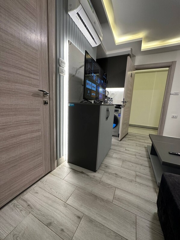 A modern hotel room with a wooden door, a minibar, a flat-screen TV, and a ceiling light fixture.