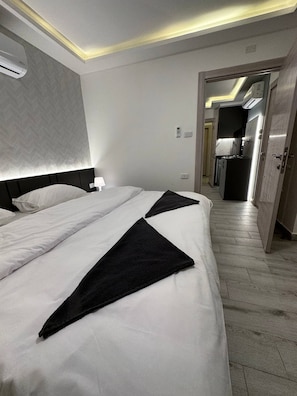 A hotel room with a large bed, a bedside table, a lamp, a wall-mounted air conditioner, a small bedside phone, and a view into the adjacent bathroom.