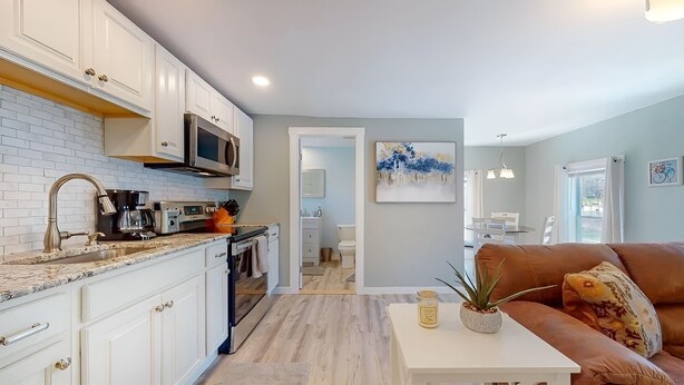 A modern kitchen with white cabinets, a microwave, and a coffee machine. There is a dining area with a brown leather couch and a white table. - Cozy Waterfall Home | Close to All Mountains/Hikes (East Durham)