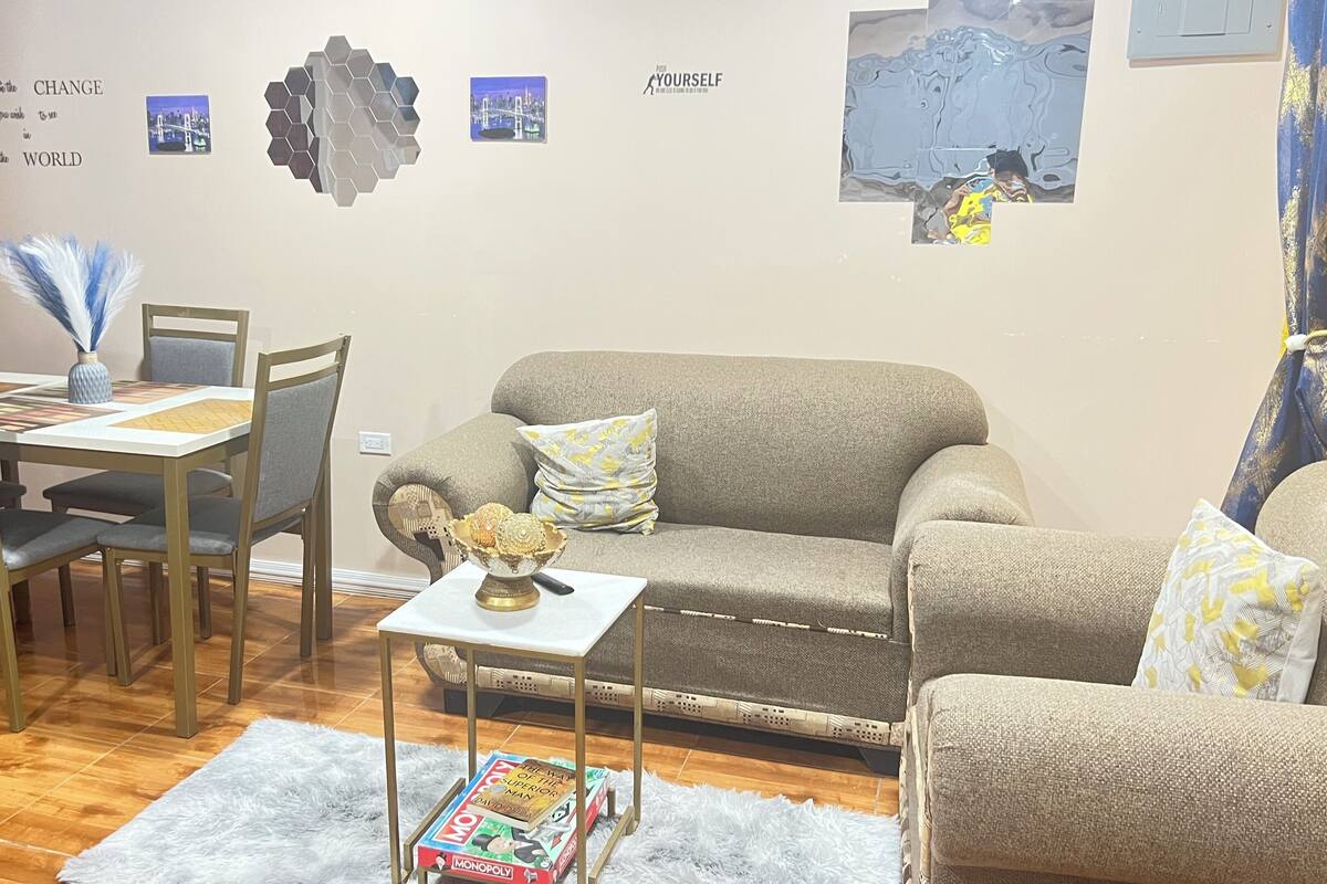 Un salon avec un canapé gris, une table à manger avec des chaises, une petite table d’appoint avec un bol, et un jeu de société Monopoly.
