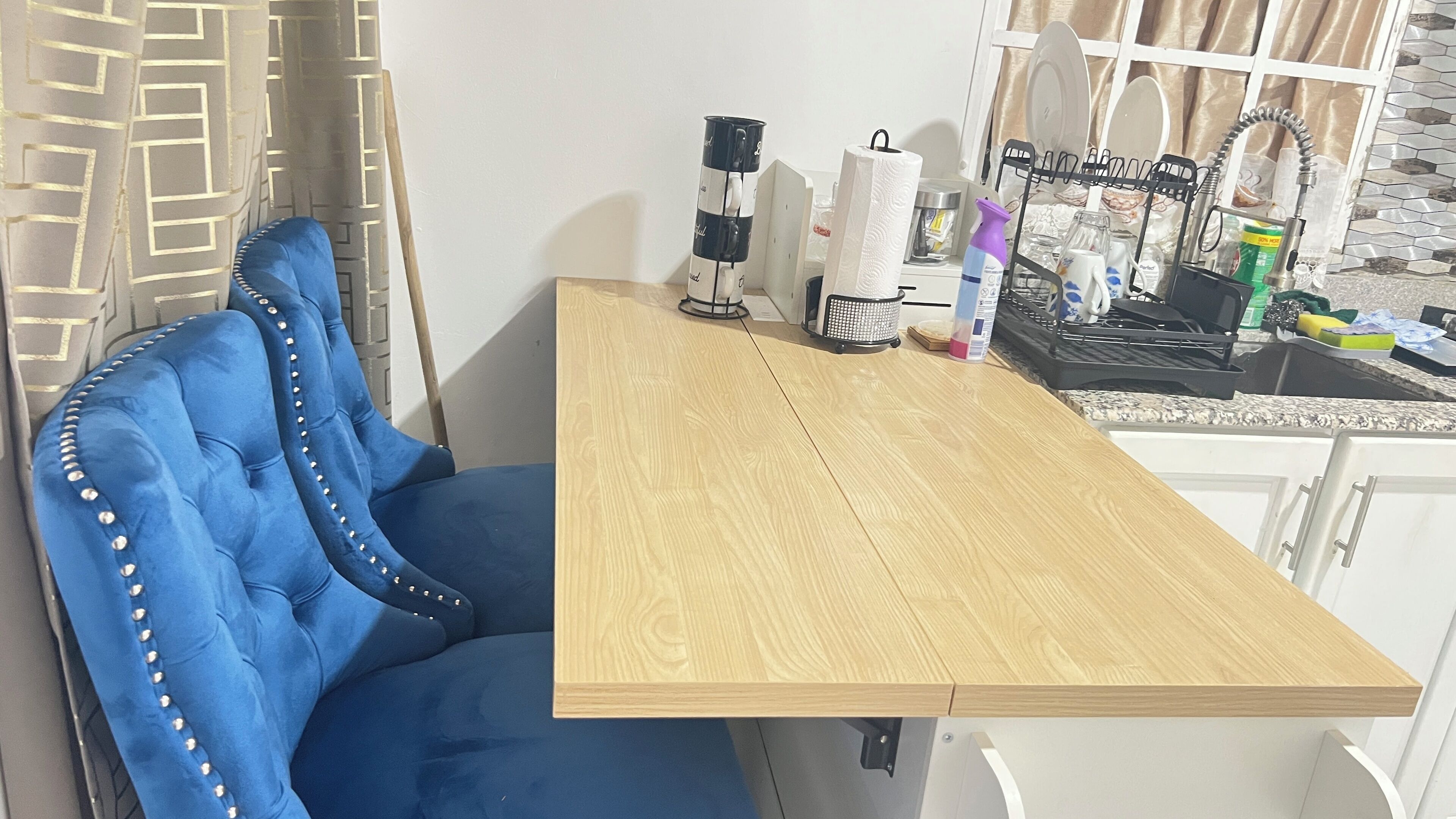 A small kitchen area with a wooden table, a blue upholstered chair, a coffee maker, and various kitchen items.