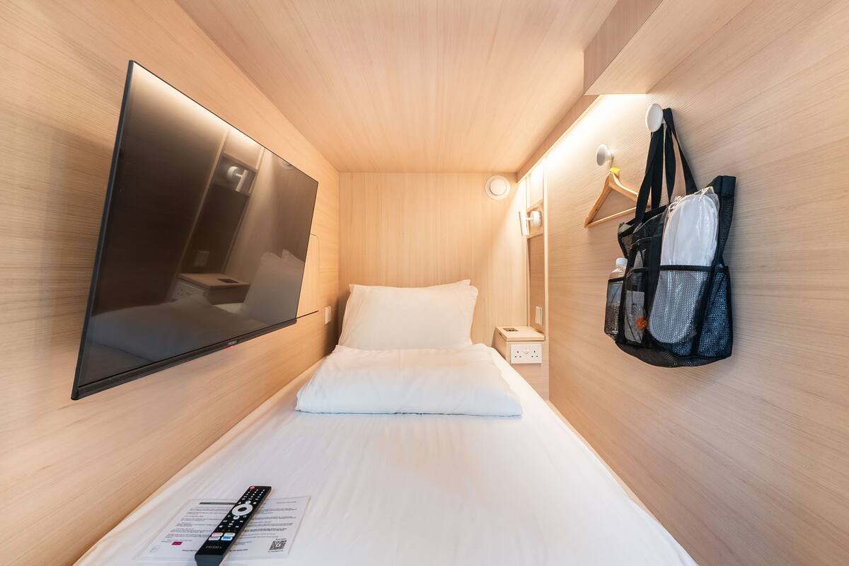 A compact hotel room with a bed, a mounted TV, a bedside table, and a wall-mounted bag rack.