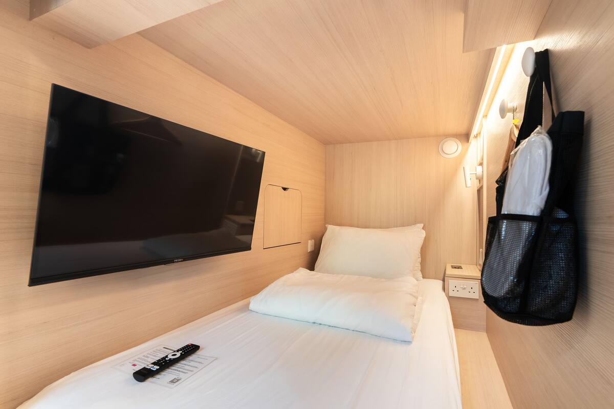 A hotel room with a bed, a TV mounted on the wall, a bedside table, and a hanging bag.