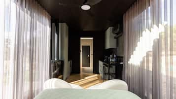 A bedroom with a bed, a ceiling fan, and a view of a kitchen area through sheer curtains.